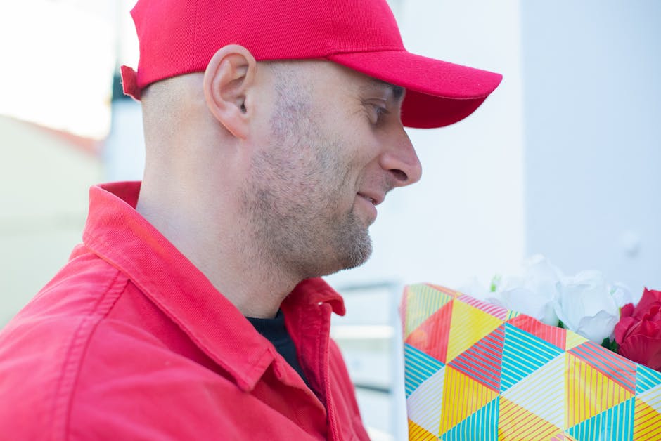 A delivery person wearing a red cap and matching red jacket is holding a colorful, geometric-patterned gift bag filled with white and pink flowers, ready for flower delivery. The focus is on the side