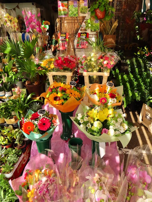 A display of various fresh flower bouquets arranged on tall green stands and white baskets at a florist shop. The bouquets include bright yellow sunflowers, pink gerbera daisies, red and white roses,