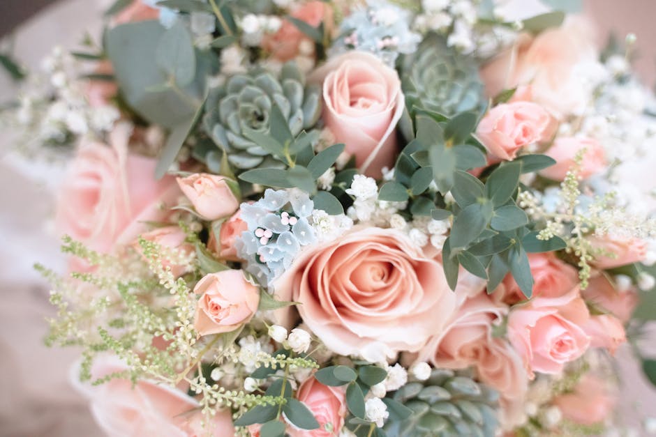 A floral arrangement featuring a bouquet of soft pink roses, complemented by eucalyptus leaves and small white flowers, arranged in a rounded, natural style. The roses have tightly furled petals and a