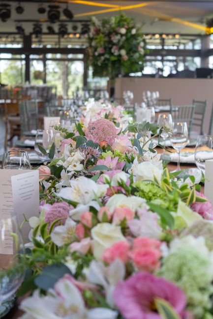 A long floral arrangement featuring a variety of fresh flowers in soft pastel shades, including pale pink roses, white lilies, light purple hydrangeas, and green foliage, is centrally placed along a t