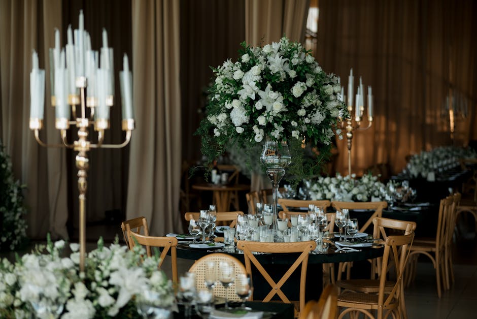 A round banquet table set for a formal event, decorated with a large, lush floral arrangement featuring white flowers such as roses, hydrangeas, and orchids, accented with greenery, and placed on a ta