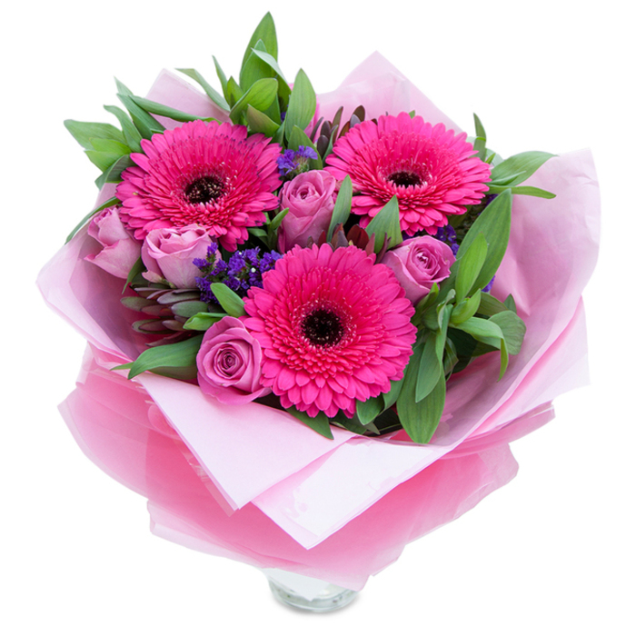 I see this hand-tied arrangement as a study in playful elegance: three wide-open gerberas in a saturated fuchsia shade present their neat concentric petals and dark centres like bright, cheerful faces, while a scattering of lavender-pink roses, still tightly budded, unfurl with layered grace and a soft, velvety texture. Small sprigs of purple statice weave through the bouquet, their papery clusters catching light and adding a whisper of cool colour that balances the warm pinks. The greenery - broad, glossy leaves with the occasional rounded eucalyptus silhouette - frames each bloom and adds a crisp, garden-fresh aroma that mingles subtly with the floral sweetness. The bouquet is lovingly cocooned: an inner sheet of pale, matte pink tissue paper forms a gentle bed, and over that a translucent fuchsia-pink cellophane, slightly crinkled and reflective, wraps the stems with an artisan's neat folds, creating a polished presentation. Near the bottom the clear glass vase rim peeks out, suggesting this is a display-ready piece intended to sit on a bedside table or kitchen counter. Picture this delivered after a morning stroll past Enfield Lock or left on a doorstep near Ponders End - it reads as an intimate embrace, ideal for marking a birthday, sending warm Mother's Day wishes, or simply telling someone they are cherished. The overall energy is bright, tender and made with the careful hands of a local florist who understands the small ways flowers lift everyday moments.